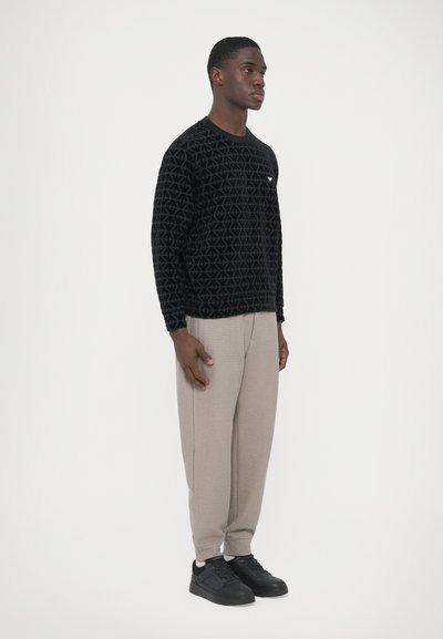 Čierna mikina s geometrickým textúrovým vzorom, svetlošedé nohavice s waffle vzorom a čierne tenisky. Model stojí bočným profilom na bielom pozadí.