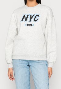 Sudadera gris clara con "NYC" impreso en negro y acentos azules, puños acanalados y un cuello redondo. Combinada con jeans azul claro.