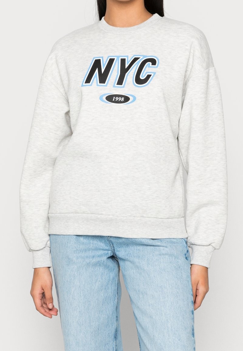 Sudadera gris clara con "NYC" impreso en negro y acentos azules, puños acanalados y un cuello redondo. Combinada con jeans azul claro.