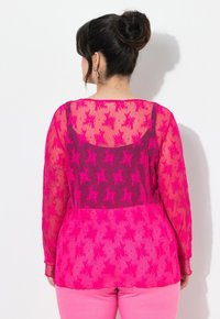 Durchsichtige rosa Bluse mit floralem Muster, langen Ärmeln und einem einfarbigen dunkelpinken Panel auf der Rückseite. Kombiniert mit rosa Hosen.