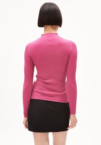 Kvinna med rakt bob-hårklipp, iklädd en åtsittande rosa ribbad långärmad tröja och svart kjol, sedd bakifrån mot en vit bakgrund.