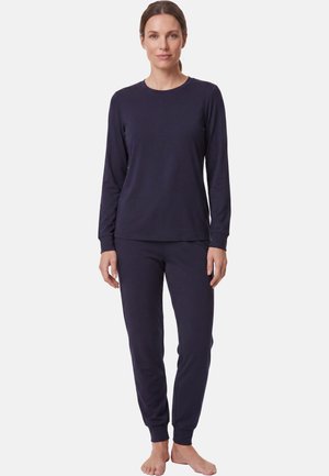 Donna in piedi a piedi nudi che indossa una maglia a maniche lunghe e pantaloni blu scuro coordinati su uno sfondo chiaro e uniforme.