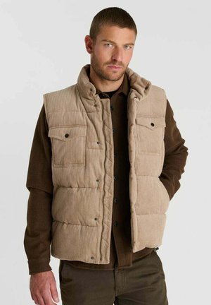 Man draagt een beige corduroy puffer-vest over een bruin shirt met lange mouwen, met één hand in de zak van de vest, kijkt vooruit.