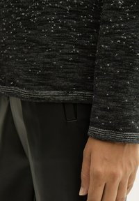 Schwarzer strukturierter Pullover mit einem gesprenkelten Muster, der einen kontrastierenden Ausschnitt und Bündchen aufweist. Eine Hand liegt im Vordergrund auf dunklen Hosen.