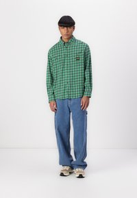 Green checkered shirt with long sleeves, buttoned front, and a black logo patch; paired with loose blue jeans and beige sneakers.