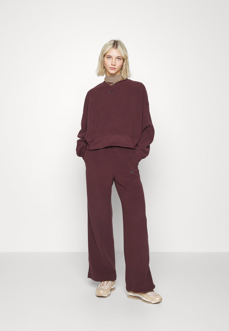 Pull en polaire bordeaux avec des épaules tombantes et un logo noir, associé à un pantalon large assorti et des baskets métalliques.
