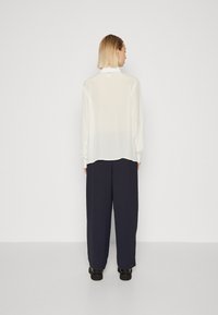 Blusa bianca a maniche lunghe con polsini arricciati, abbinata a pantaloni navy larghi e calzature nere; tessuto morbido e leggero.