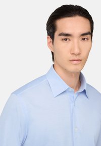 Ung man med mörkt hår iförd en ljusblå skjorta med knappar, som tittar rakt fram mot en enkel vit bakgrund.