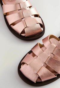 Rosa metalliska flätade sandaler med en platt, brun sula och breda remmar. Texturerat material skapar en lager-på-lager, kors och tvär design över ovansidan.
