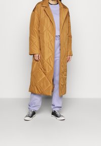 Dlouhý prošívaný kabát v zlatohnědé barvě s ostrým límcem, spárovaný s levandulovým sweatšortem a černými teniskami. Texturovaná, lehká tkanina.