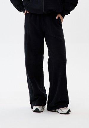 Osoba w luźnych czarnych spodniach i białych tenisówkach, stojąca z rękami w kieszeniach na jednolitym tle.