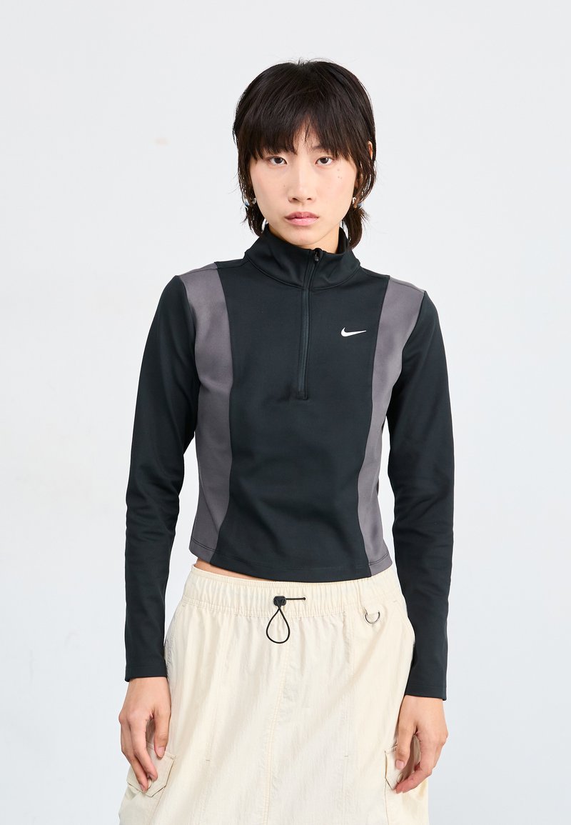 Top negru cu mânecă lungă, cu fermoar, cu panouri laterale gri și guler fals; prezintă un mic logo Nike pe piept și un design ajustat.