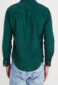 Camicia verde scuro con bottoni, orlo curvo, particolare del giogo appuntito e maniche lunghe arrotolate ai polsini. Realizzata in tessuto strutturato.