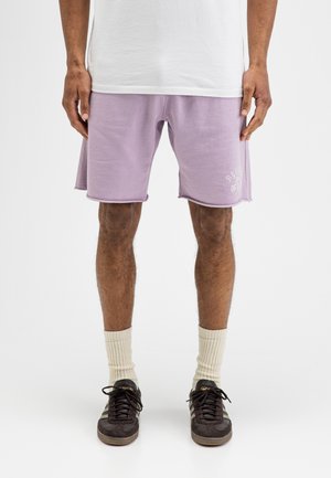 Mann trägt hellviolette Shorts, weißes Hemd, cremefarbene Socken und schwarze Turnschuhe, steht vor einem schlichten weißen Hintergrund.