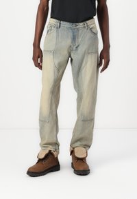 Jeans din denim de culoare albastră deschisă, cu o croială relaxată, având buzunare unghiulare și detalii vizibile de cusătură, asortați cu botine maro cu șireturi.