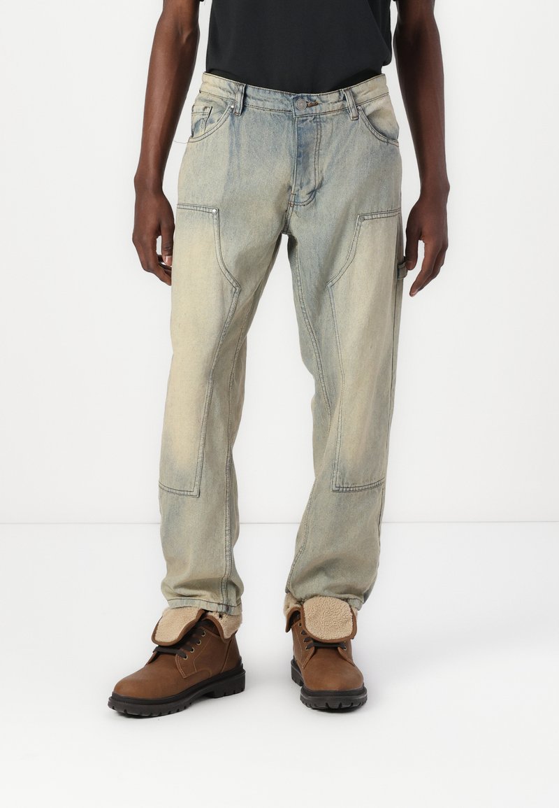 Jeans din denim de culoare albastră deschisă, cu o croială relaxată, având buzunare unghiulare și detalii vizibile de cusătură, asortați cu botine maro cu șireturi.