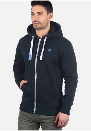 Veste à capuche zippée navy avec cordons blancs, poches avant et logo brodé. Tissu doux et poignets côtelés. Détail de fermeture éclair argentée.