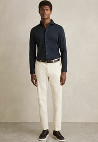Cămașă bleu cu mânecă lungă, pantaloni de culoare Crem, adidași închis. Prezintă un design cu nasturi, o croială ajustată și accente din curea de piele.