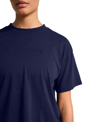 Vrouw draagt een marineblauw T-shirt met "HALO" geborduurd op de linkerborst, afgebeeld van schouders tot midden van de torso tegen een witte achtergrond.