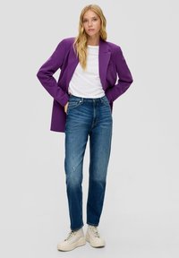 Fioletowa marynarka na białej koszulce, w połączeniu z niebieskimi przetartymi dżinsami i jasnobeżowymi sneakerami. Minimalna biżuteria, neutralne tło.