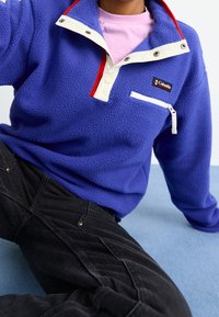 Pullover in pile viola con colletto a scatto bianco e rosso, tasca con zip sul petto e logo Columbia. Abbinato a pantaloni neri con texture.