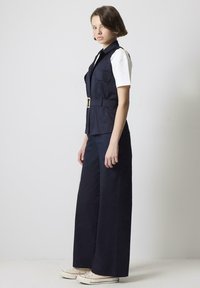 Námornícky vest s opaskom v páse a dvoma prednými vreckami, skombinovaný s širokými námorníckymi nohavicami a bielymi teniskami. Model stojí v profile.