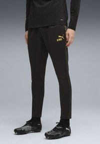 Svarta träningsbyxor med guld "KING"-logotyp, med en slim fit och strukturerat sidofin mönster, tillsammans med svarta fotbollsskor.