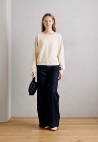 Felpa crema con testo in rilievo, abbinata a jeans neri a vita alta e una borsa nera strutturata. Modello con scarpe di colore chiaro.