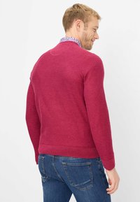 Gestrickter Pullover in sattem Magenta, mit langen Ärmeln, rundem Halsausschnitt, geripptem Saum und strukturierter Oberfläche. Über einem floralen Hemd mit Kragen getragen.