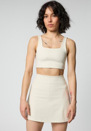Kvinde med kort krøllet hår iført en cremefarvet crop top og matchende højtaljet nederdel, der viser tatoveringer på brystet og underarmen.
