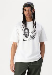 White cotton t-shirt featuring a graphic print of a face and text on the front. Short sleeves and crew neckline, paired with olive green pants.