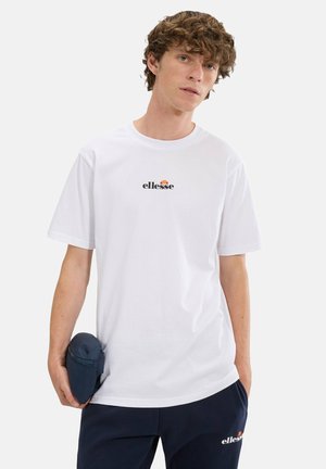 Junger Mann, der ein weißes Ellesse-T-Shirt und marineblaue Hose trägt, hält eine kleine marineblaue Tasche in einer Hand und steht vor einem einfarbigen Hintergrund.