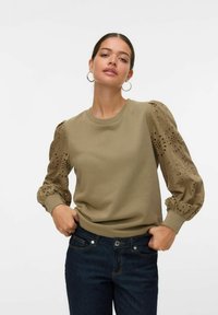 Sudadera verde oliva con mangas abullonadas texturizadas que presentan detalles de ojales. Cuello redondo clásico y puños acanalados. Combinada con jeans oscuros.