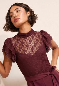 Top de encaje burdeos con un cuello alto, panel de encaje floral transparente y mangas con volantes. Combinado con un detalle de lazo en la cintura a juego.
