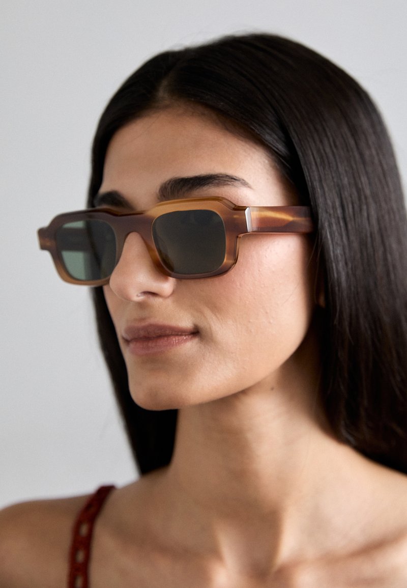 Ochelari de soare dreptunghiulari, de culoare maronie, cu un cadru gros, transparent și lentile întunecate. Un model subtil de marmură este vizibil pe suprafața cadrului.