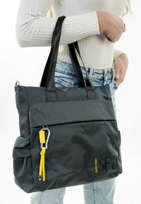 Black textured tote bag with black handles and a zippered pocket, featuring a yellow accent and logo. Measures medium size.