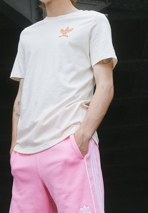 Personne portant un T-shirt Adidas blanc avec logo orange et un short Adidas rose avec bandes blanches, mains dans les poches, tatouages visibles sur les bras.