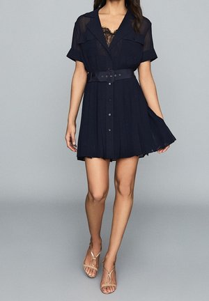 Kobieta ubrana w granatową sukienkę z krótkim rękawem i paskiem, ozdobioną koronkowymi detalami, w połączeniu z beżowymi sandałami na obcasie z odkrytymi palcami, stojąca na szarym tle.