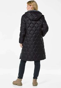 Schwarze gesteppte Jacke mit Kapuze, die ein strukturiertes, gepolstertes Design aufweist. Getragen über dunklen Jeans und hellbraunen Stiefeln, mit einem mittellangen Stil.