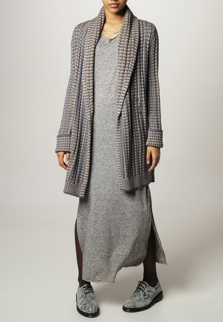Femme portant une longue robe grise, un cardigan à motifs ouvert sur le devant, des collants noirs et des chaussures à lacets gris en cuir effet peau de serpent.