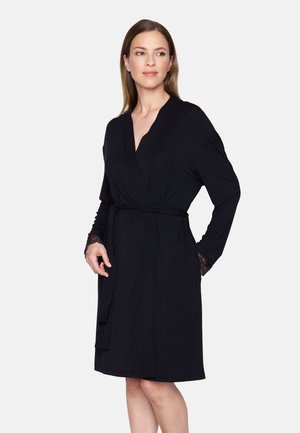 Mujer con un vestido envolvente negro hasta la rodilla, con mangas acabadas en encaje, posando con una mano en el bolsillo contra un fondo liso.