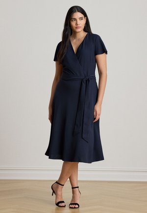 Une femme porte une robe portefeuille bleu marine avec un lien à la taille et des talons noirs, devant un fond de studio uni.