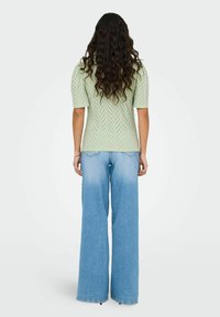 Lys grøn, kortærmet strikket top med chevron mønster, parret med vide ben, lyse jeans, set fra bag.