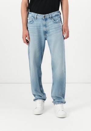Hombre con vaqueros sueltos de color azul claro combinados con zapatillas blancas y una camisa negra, de pie frente a un fondo blanco liso.