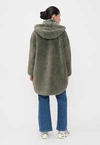 Överdriven, olivgrön fleecejacka med huva, mjuk textur och rundad kant, tillsammans med utsvängda blå jeans och vita sneakers.