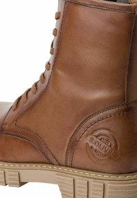 Botín de cuero marrón con puntera redonda, diseño de cordones y suela de goma texturizada con detalle de logo en relieve.