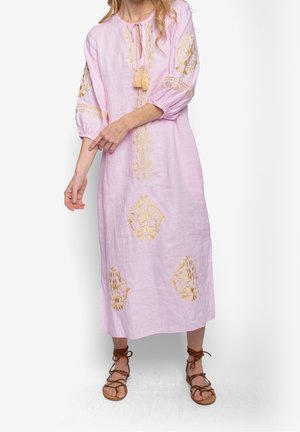 Femme portant une robe midi rose pâle avec broderie dorée, manches trois-quarts, encolure nouée avec petits pompons, et sandales marron à lacets.