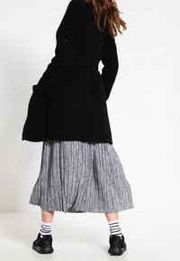 Žena oblečená v černém dlouhém svetry, šedé skládací sukni, černých teniskách a bílých ponožkách s černými pruhy, stojící na bílém pozadí.