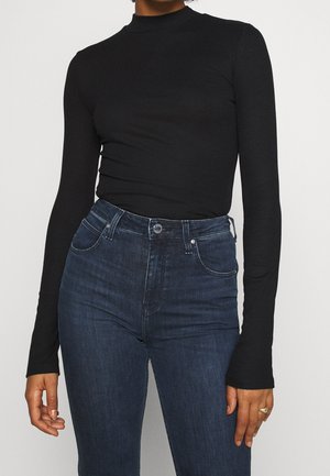 Personne portant un haut noir ajusté à manches longues, rentré dans un jean taille haute bleu foncé, debout devant un fond uni.