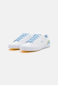 White sneakers with blue laces, featuring a low-top design, smooth texture, and a side yellow and blue accent detail. Rubber sole.